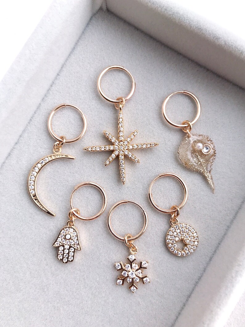 May include: Six gold hoop earrings with different charms. The charms include a crescent moon, a star, a seashell, a hamsa hand, a snowflake, and a circle.