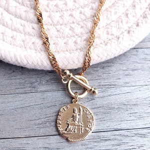May include: A gold-toned necklace featuring a twisted chain and a toggle clasp. A circular pendant hangs from the clasp, displaying a relief of a seated figure and the text "IVPITER CUSTOS" embossed along the edge of the coin-shaped charm.