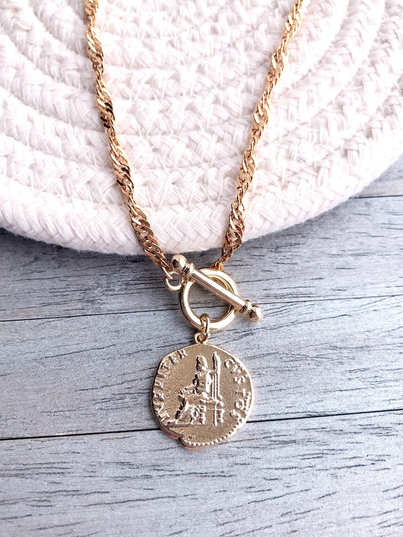May include: A gold-toned necklace featuring a twisted chain and a toggle clasp. A circular pendant hangs from the clasp, displaying a relief of a seated figure and the text "IVPITER CUSTOS" embossed along the edge of the coin-shaped charm.