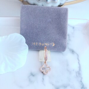 Puede incluir: Aretes de aro de oro rosa con un dije colgante de tr&eacute;bol de cuatro hojas, ambos adornados con piedras de circonita c&uacute;bica transparentes. Los aretes se muestran en una bolsa de terciopelo gris con el texto "ME GUMI" impreso en ella.