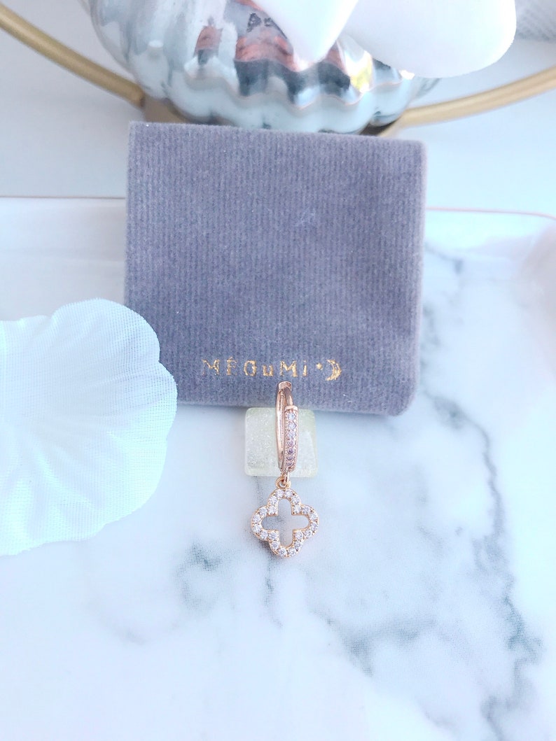 Puede incluir: Aretes de aro de oro rosa con un dije colgante de tr&eacute;bol de cuatro hojas, ambos adornados con piedras de circonita c&uacute;bica transparentes. Los aretes se muestran en una bolsa de terciopelo gris con el texto "ME GUMI" impreso en ella.