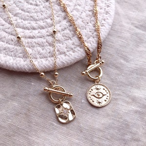 Op de afbeelding: Drie gouden kettingen met verschillende kettingstijlen en bedels. Eén ketting heeft een kleine sterbedel met strass, een andere heeft een ronde bedel met een oog en sterren, en de derde heeft een schuifgesp.