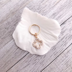 May include: A gold hoop earring with a dangling charm in the shape of a four-leaf clover. The clover is encrusted with clear rhinestones.