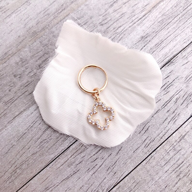 May include: A gold hoop earring with a dangling charm in the shape of a four-leaf clover. The clover is encrusted with clear rhinestones.
