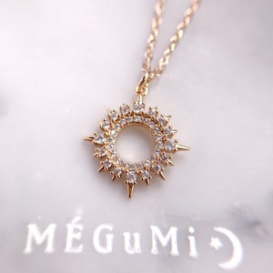 May include: Gold necklace with a sun pendant featuring a circle of small clear stones and a larger circle of clear stones around the edge. The sun pendant has points radiating out from the circle.