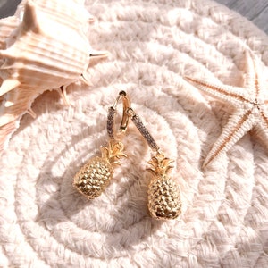May include: A pair of gold hoop earrings with a pineapple charm dangling from each hoop. The earrings are on a white textured surface.