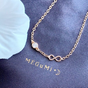 Könnte beinhalten: Ein goldenes Kettenarmband mit einem Unendlichkeits-Symbol-Anhänger und einem einzigen klaren Edelstein. Das Armband liegt auf einem schwarzen Stoffhintergrund mit dem Text "MEGUMI" in goldenen Buchstaben.