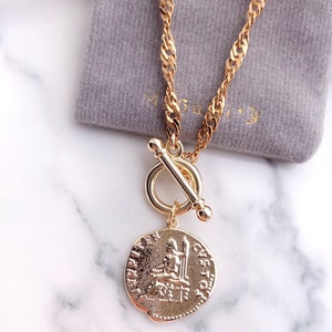May include: A gold-toned necklace featuring a twisted chain and a toggle clasp closure. A circular pendant hangs from the toggle, embossed with a classical figure seated on a throne and the text JUPITER and CASTOR along the outer edge.
