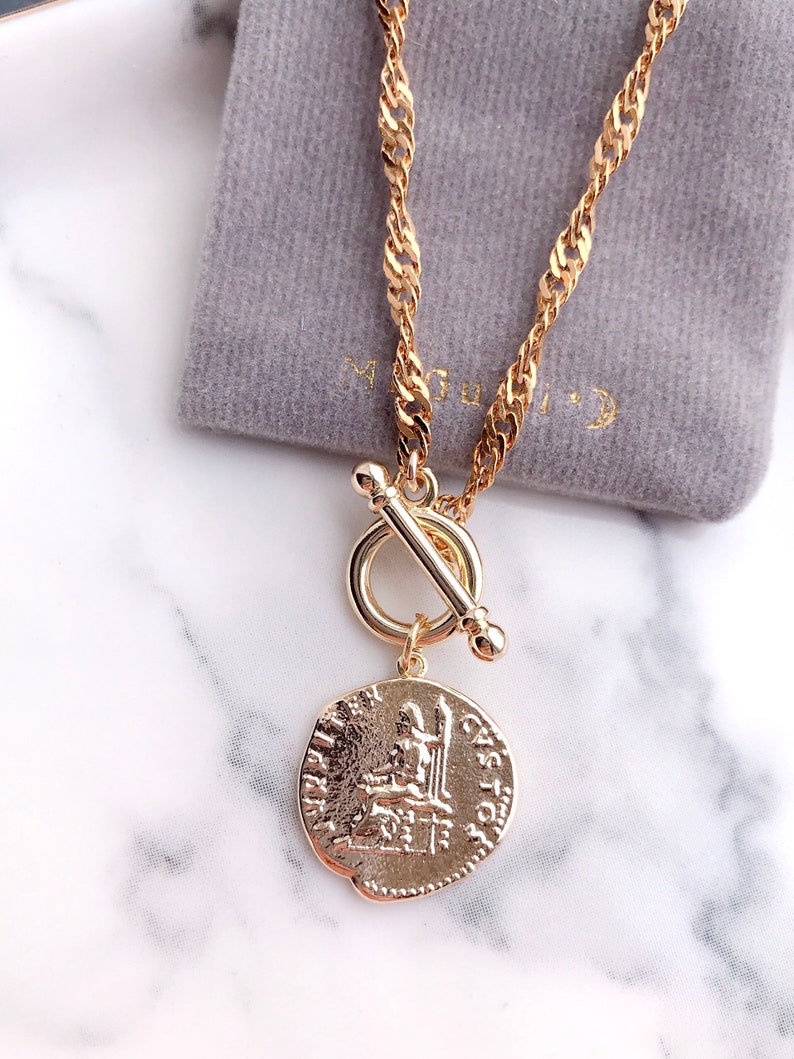 May include: A gold-toned necklace featuring a twisted chain and a toggle clasp closure. A circular pendant hangs from the toggle, embossed with a classical figure seated on a throne and the text JUPITER and CASTOR along the outer edge.