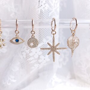 May include: Six gold hoop earrings with different charms hanging from them. The charms include a snowflake, an eye, a circle, a star, a dried flower, and a small star.