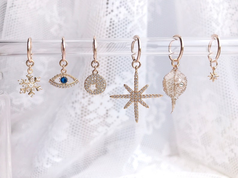 May include: Six gold hoop earrings with different charms hanging from them. The charms include a snowflake, an eye, a circle, a star, a dried flower, and a small star.