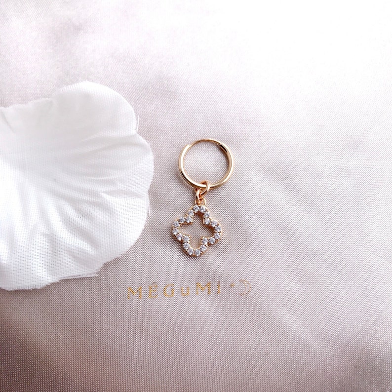 May include: Gold hoop earrings with a dangling charm in the shape of a four-leaf clover, adorned with clear cubic zirconia stones.