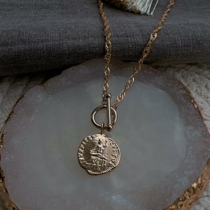 May include: A gold-toned necklace featuring a twisted chain and a toggle clasp closure. A circular pendant hangs from the toggle, displaying an embossed classical figure on its surface with intricate detailing and lettering around the edge.