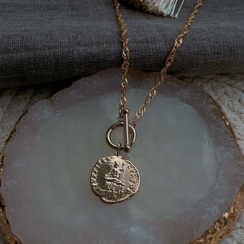 May include: A gold-toned necklace featuring a twisted chain and a toggle clasp closure. A circular pendant hangs from the toggle, displaying an embossed classical figure on its surface with intricate detailing and lettering around the edge.