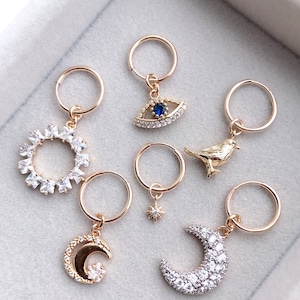 May include: Set of six gold hoop earrings with different charms. The charms include a sun, a moon, a crescent moon, a bird, a star, and an evil eye. Each charm is adorned with clear rhinestones.