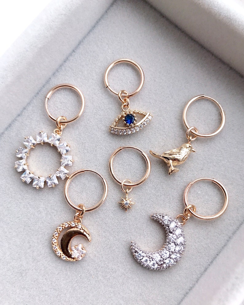 May include: Set of six gold hoop earrings with different charms. The charms include a sun, a moon, a crescent moon, a bird, a star, and an evil eye. Each charm is adorned with clear rhinestones.