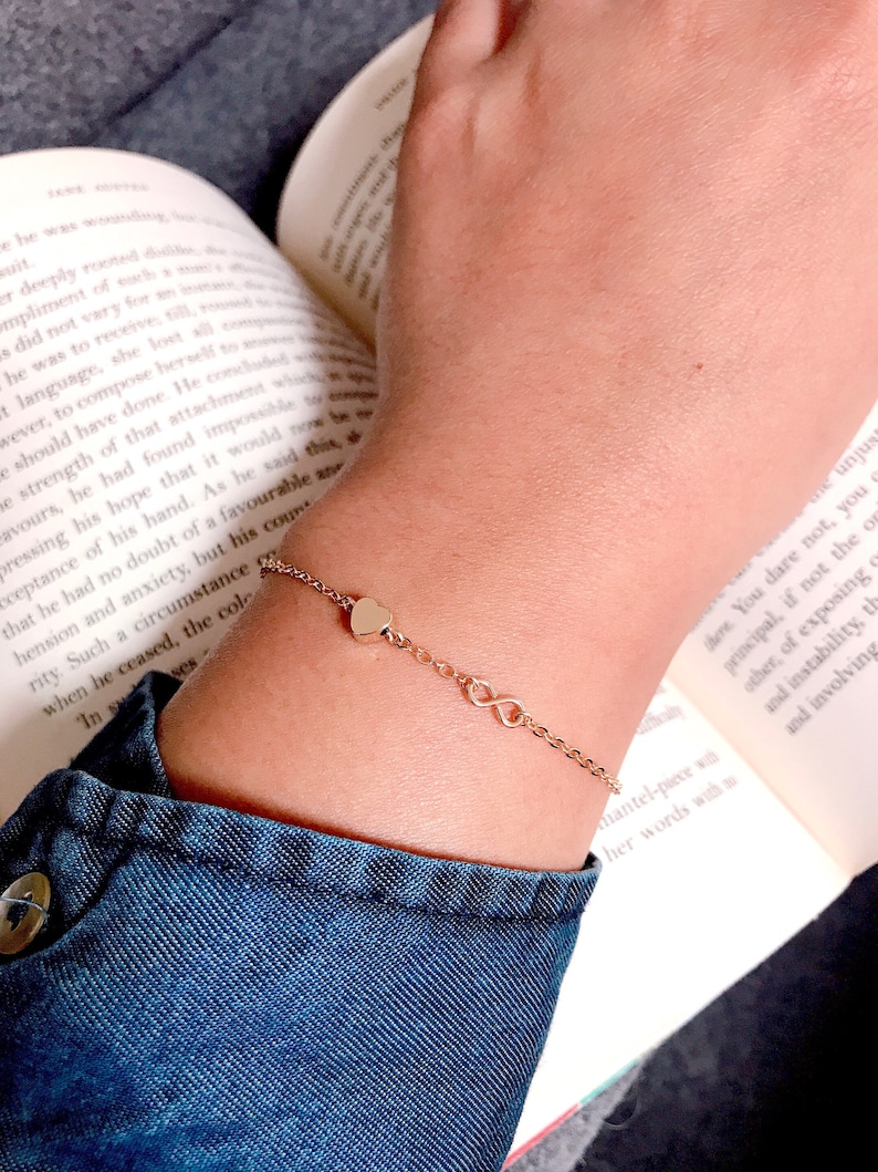 May include: A gold chain bracelet with a heart and infinity symbol charms. The bracelet is on a person's wrist.