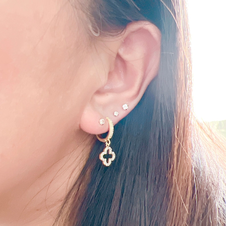 Puede incluir: Aretes de aro dorados con un dije colgante de tr&eacute;bol de cuatro hojas. El tr&eacute;bol est&aacute; incrustado con peque&ntilde;as piedras transparentes.
