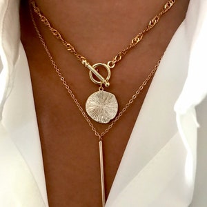 May include: A layered gold-tone necklace set featuring a top chain with a toggle clasp and a round textured pendant, paired with a lower delicate chain featuring a long, thin vertical bar pendant, worn over a crisp white collared shirt.