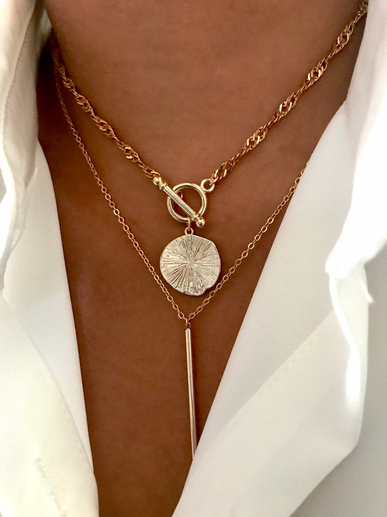 May include: A layered gold-tone necklace set featuring a top chain with a toggle clasp and a round textured pendant, paired with a lower delicate chain featuring a long, thin vertical bar pendant, worn over a crisp white collared shirt.