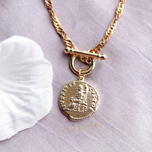May include: A gold-toned necklace featuring a twisted chain and a toggle clasp. A circular pendant hangs from the clasp, embossed with a figure seated on a throne and the text JUPITER CUSTOS along the edge. The metal has a polished finish.