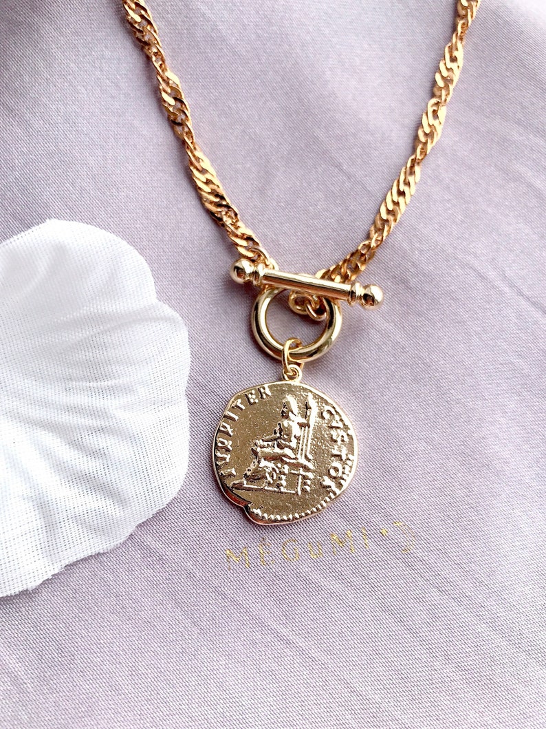 May include: A gold-toned necklace featuring a twisted chain and a toggle clasp. A circular pendant hangs from the clasp, embossed with a figure seated on a throne and the text JUPITER CUSTOS along the edge. The metal has a polished finish.