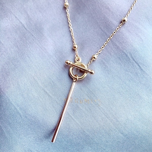 Könnte beinhalten: Eine goldene Halskette mit einem Toggle-Verschluss und einem langen, dünnen Anhänger. Die Halskette liegt auf einem blauen Stoffhintergrund.