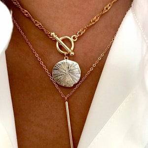 May include: A layered gold-tone necklace set featuring a top chain with a twisted link design and a toggle clasp. The second chain includes a round, textured pendant with radiating lines and a long, smooth, vertical bar pendant hanging below.