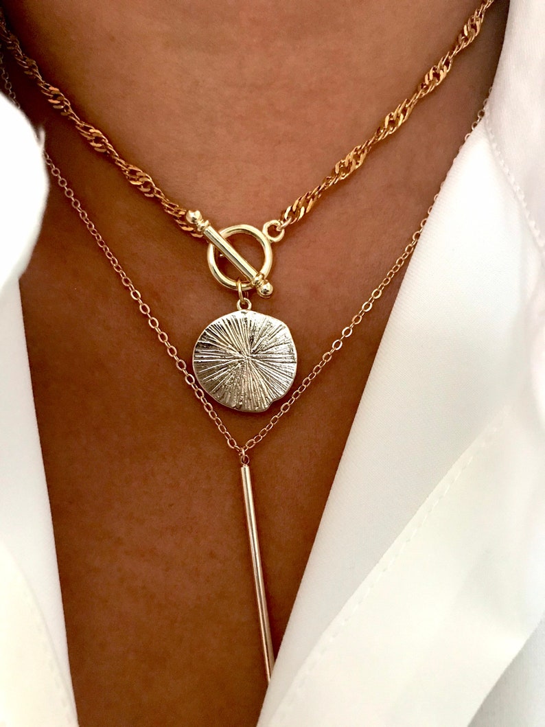 May include: A layered gold-tone necklace set featuring a top chain with a twisted link design and a toggle clasp. The second chain includes a round, textured pendant with radiating lines and a long, smooth, vertical bar pendant hanging below.
