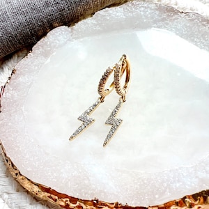 May include: A pair of gold hoop earrings with a dangling lightning bolt charm. Each charm is encrusted with small, clear cubic zirconia stones.