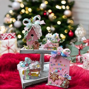 May include: A festive display of Christmas-themed decorations. A pink birdhouse, a gift box, and a sign that reads "Gingerbread Bakery" are adorned with bows and gingerbread men. The items are set on a white tray against a backdrop of a Christmas tree.