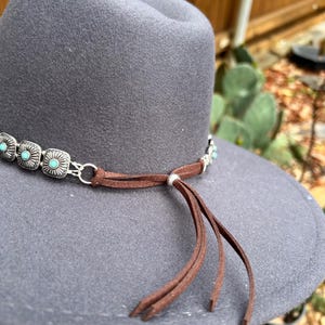 Puede incluir: Sombrero de fieltro gris con una banda decorativa con piedras de color turquesa y detalles met&aacute;licos plateados. Una banda de ante marr&oacute;n con borlas est&aacute; atada alrededor del sombrero. Estilo occidental.
