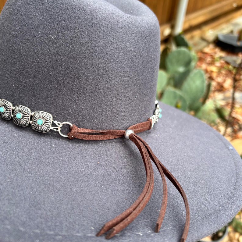 Puede incluir: Sombrero de fieltro gris con una banda decorativa con piedras de color turquesa y detalles met&aacute;licos plateados. Una banda de ante marr&oacute;n con borlas est&aacute; atada alrededor del sombrero. Estilo occidental.