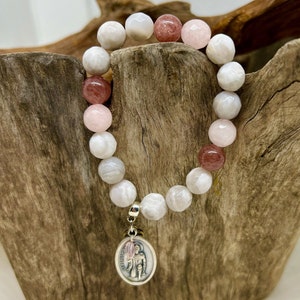 Puede incluir: Una pulsera de cuentas con cuentas de piedras preciosas blancas, rosas y marrón rojizo. Un colgante plateado con la imagen de un santo cuelga de la pulsera. La pulsera se exhibe sobre un trozo de madera. Las cuentas miden aproximadamente 1 cm de diámetro.