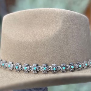 May include: A tan felt cowboy hat with a silver and turquoise hat band. The hat band is made of a repeating pattern of silver crosses with turquoise stones in the center.