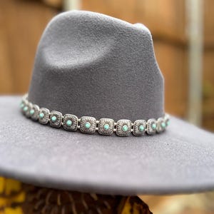 Puede incluir: Un sombrero gris estilo fedora con una banda decorativa. La banda es plateada con detalles cuadrados, cada uno con un centro de color turquesa. El sombrero tiene un ala ancha y un dise&ntilde;o cl&aacute;sico de inspiraci&oacute;n occidental.