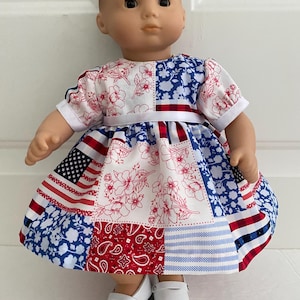May include: A doll wearing a patchwork dress with patriotic patterns. The dress features sections of the American flag, floral prints, and paisley designs in red, white, and blue. The doll is also wearing white shoes.