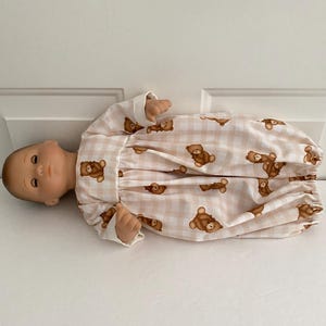 May include: A doll wearing a light pink and white gingham patterned sleep sack with brown teddy bear graphics. The sleep sack has long sleeves with white cuffs.