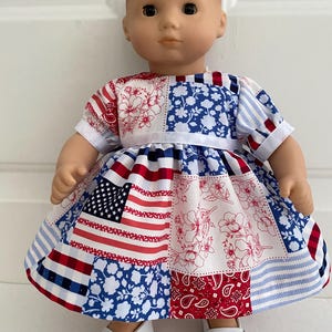 May include: A doll wearing a patchwork dress with red, white, and blue patterns, including the American flag, floral designs, and paisley. The doll is also wearing white shoes.
