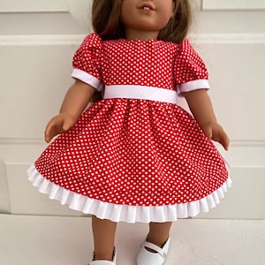 May include: A doll wearing a red dress with white heart patterns. The dress has white trim on the sleeves and hem, and a white belt. The doll is also wearing white shoes.