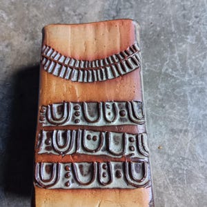 May include: A rectangular ceramic vase with a textured surface. The vase is mainly brown with decorative patterns in a darker brown and off-white. The patterns include curved and dotted designs.