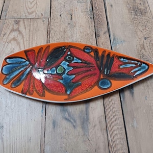 Puede incluir: Una fuente de cerámica oblonga con una base naranja vibrante. La fuente presenta un diseño abstracto audaz en rojo, negro y azul. El diseño recuerda un motivo floral o de insecto estilizado. La fuente mide aproximadamente 40 cm de largo.