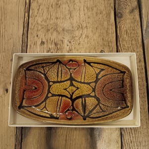 Könnte beinhalten: Ein rechteckiger Keramikservierteller mit einem abstrakten braunen, gelben und roten Design. Der Teller befindet sich in einer weißen Schachtel auf einem Holzgrund. Das Design weist geschwungene und geometrische Formen auf.