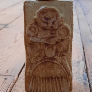 Puede incluir: Un tarro de cerámica beige rectangular con tapa. El tarro presenta un relieve tallado de una figura con cabeza redonda y detalles circulares. El tarro está sobre una superficie de madera.