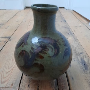 Könnte beinhalten: Eine handgefertigte Keramikvase mit rundem Körper und schmalem Hals. Die Vase ist hauptsächlich olivgrün mit dunkelbraunen abstrakten Designs. Die Vase steht auf einem Holzboden.