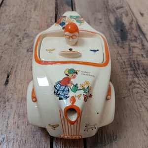 Könnte beinhalten: Cremefarbene Keramik-Teekanne mit orangefarbenen Zierleisten, mit einem Deckel mit einem gesichtsähnlichen Griff. Die Teekanne ist mit Illustrationen einer Frau, die Blumen gießt, und kleinen Vögeln verziert. Der Text "WENDY WATERING" ist sichtbar.