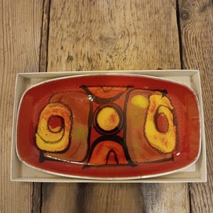 Può includere: Un piatto rettangolare in ceramica rossa con bordo bianco, con un design astratto in giallo, arancione e nero. Il piatto è all'interno di una scatola di cartone bianca, su uno sfondo di legno.