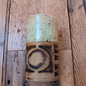 May include: A handmade ceramic mug with a two-tone design. The top half is a light green, while the bottom half is a tan color with a geometric pattern. The mug is cylindrical and appears to be hand-thrown.