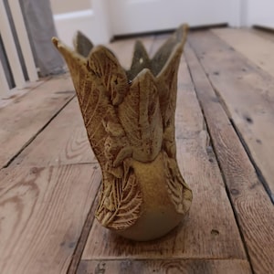 Puede incluir: Un jarrón de cerámica beige con diseño de hojas. El jarrón tiene una parte superior ancha y abierta con bordes puntiagudos y una base redondeada. La superficie del jarrón está texturizada con patrones de hojas detallados.