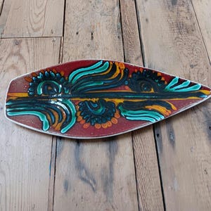 Puede incluir: Una fuente de cerámica oblonga con base roja y borde blanco. La fuente presenta un diseño central de formas abstractas turquesas, amarillas y negras. El diseño recuerda a una pluma o una hoja estilizada.